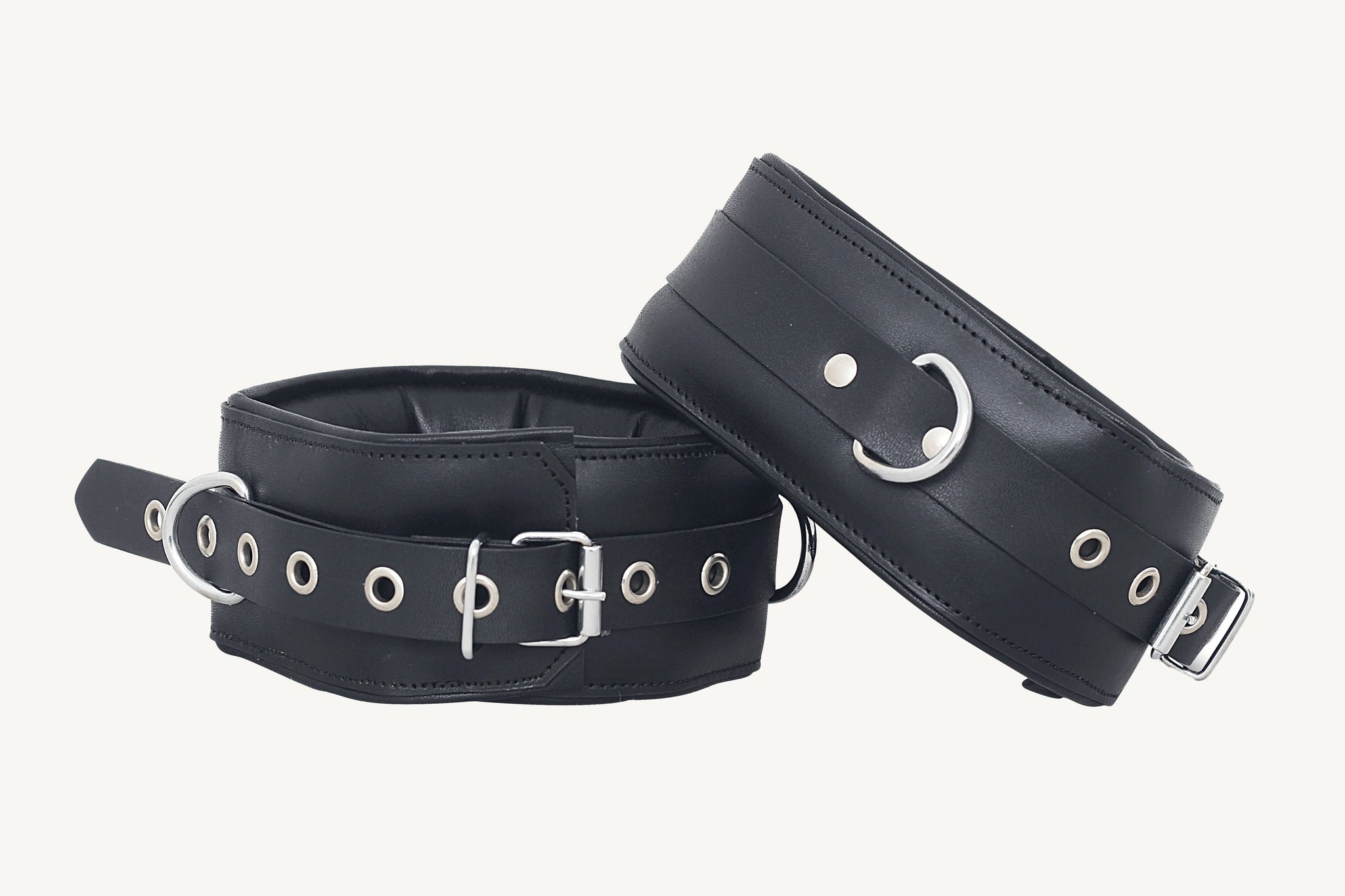 A set of black leather bondage handcuffs with metal buckles and eyelets, arranged on a white surface.