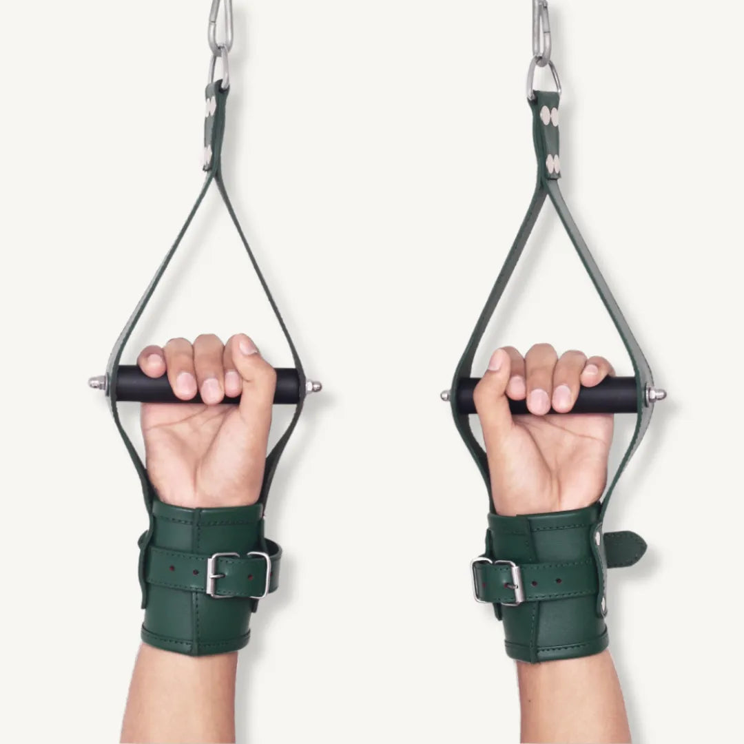 Hands facing forward gripping black rods while secured in green leather wrist suspension cuffs with straps.