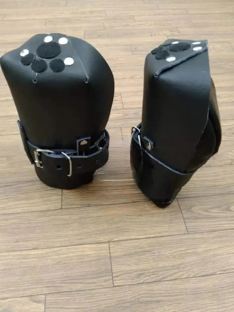 Front view of a pair of black handmade leather bondage mittens with adjustable straps, featuring circular padded designs on top, is displayed on a wooden floor.