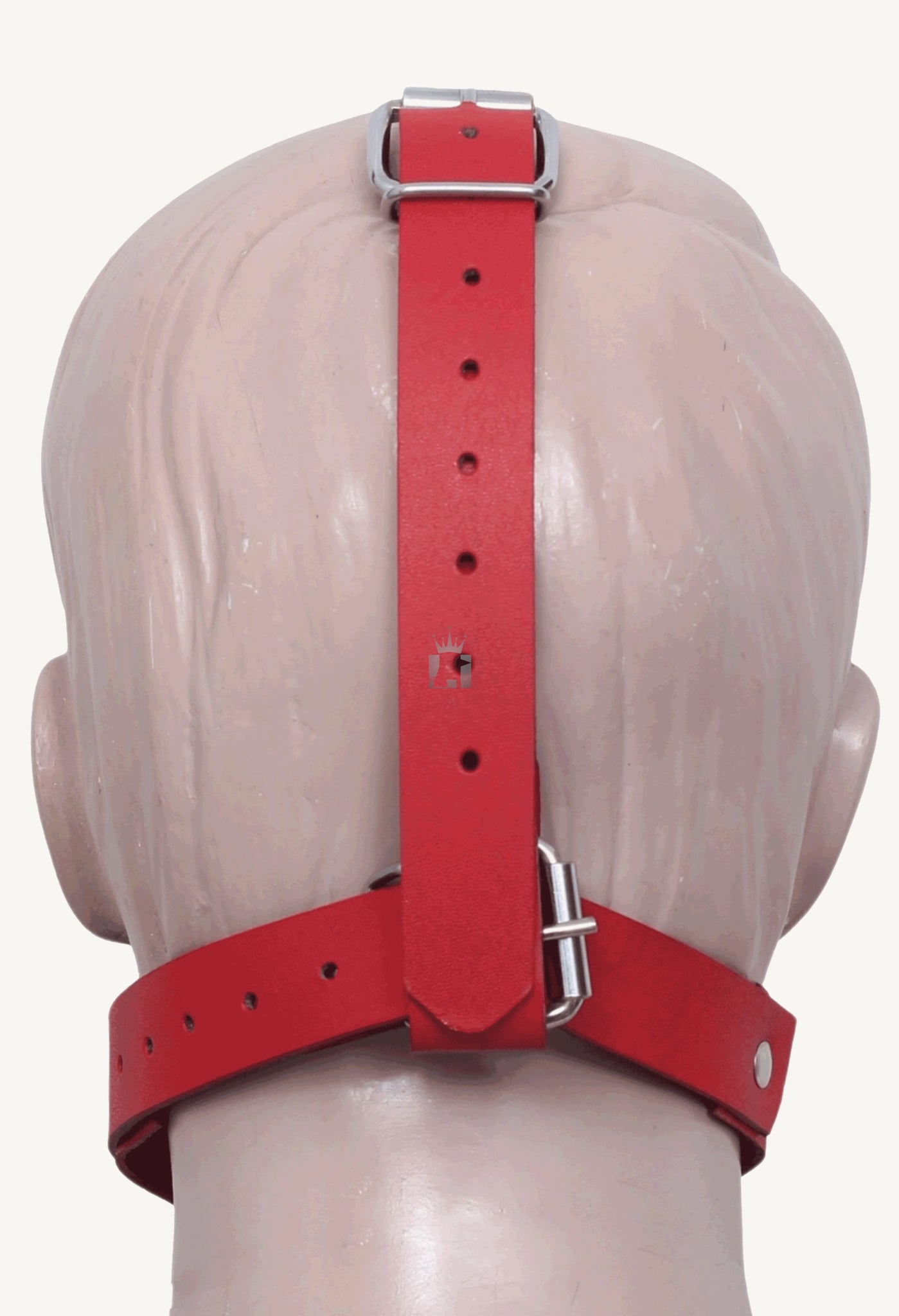 The back of a mannequin's head shows the adjustable strapping of the red leather muzzle. A vertical and a horizontal strap, both with silver buckles, secure the pet play mask in place.
