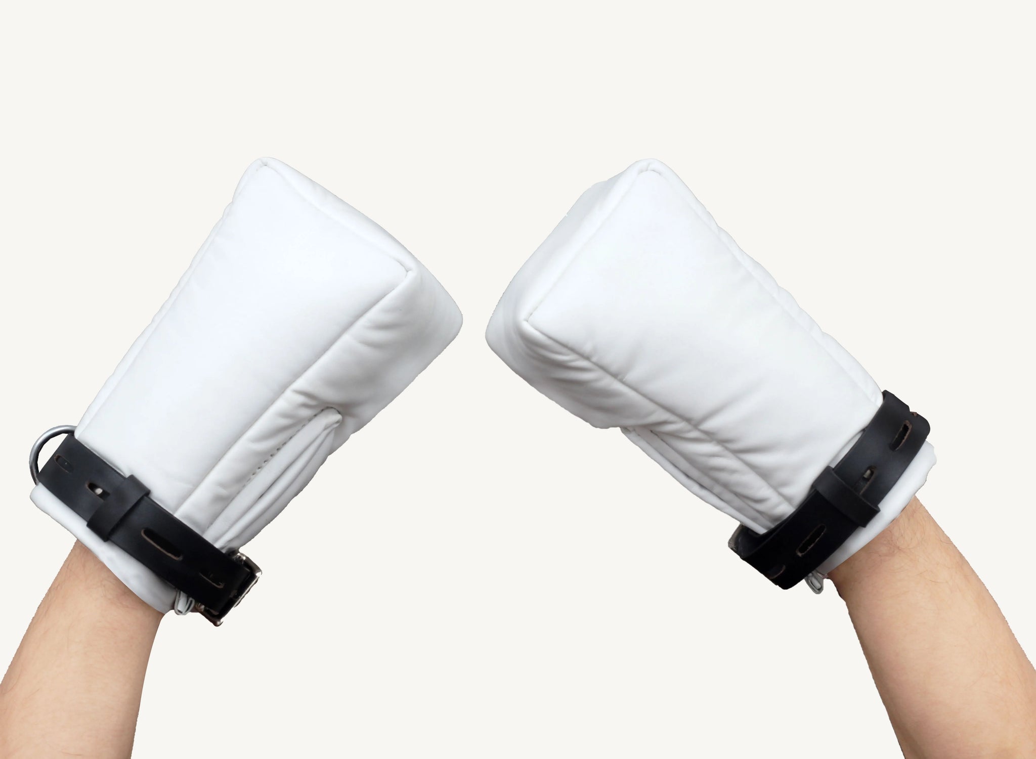 Front angle view of a hand wearing large white leather bondage mitts with black straps, raised in a playful gesture against a neutral background.